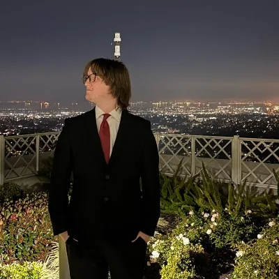 Zach in a black suit and red tie, standing on a terrace above city lights at night.